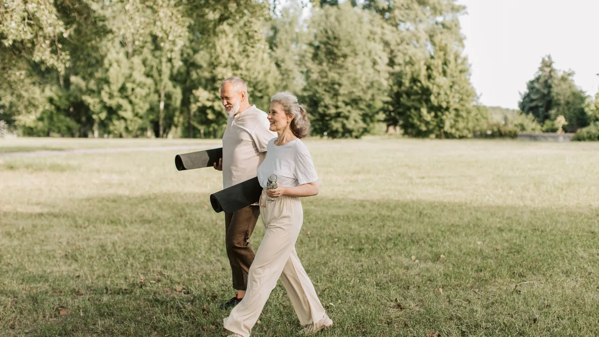 Elderly couple walking on grass carrying yoga mats, enjoying a sunny day outdoors.
