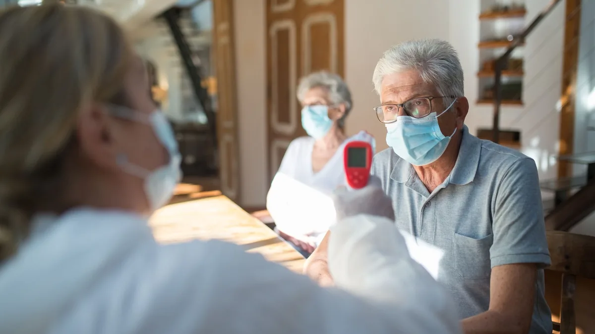 Healthcare worker checks temperature of senior adults during pandemic using a thermometer in a home setting.